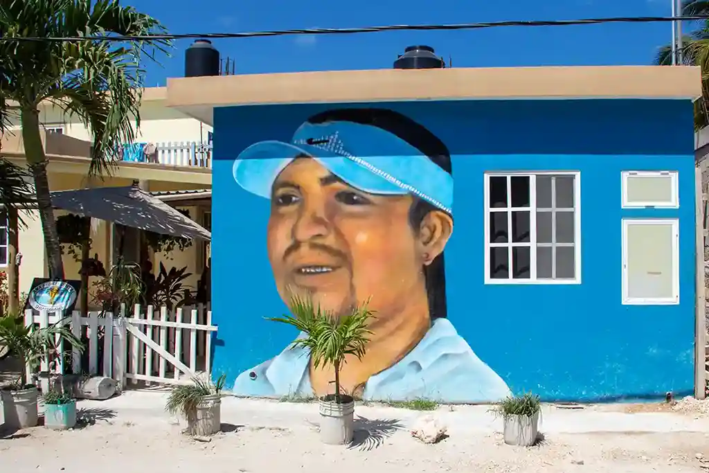 Wall painting of a man with a headband surrounded by tropical leaves on a blue house in Holbox.