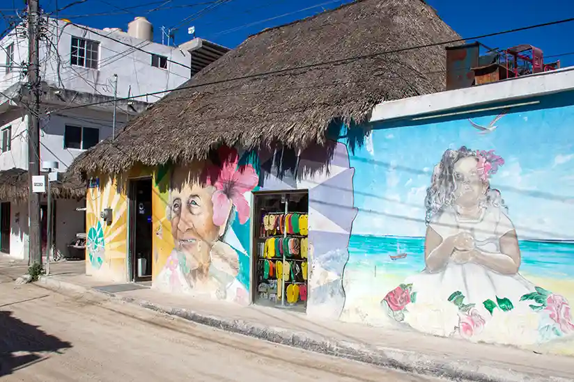 Faded and weathered mural of a girl with hibiscus flowers on a traditional thatched roof house.
