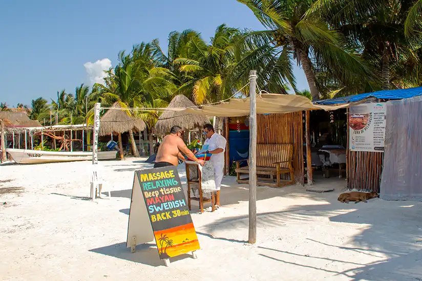 A massage stand with a sunshade and board on the beach of Isla Holbox, surrounded by palm trees.