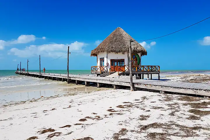 Langer Holzsteg mit einem traditionellen strohgedeckten Pfahlhaus über dem seichten Wasser von Holbox.